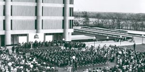 1968.073.005 Spring Convocation A large audience watches a university graduation ceremony on a rooftop next to Schmon Tower at Brock University in the 1960s inthis black and white photo.