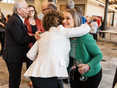 Two women wearing business attire embrace at a lively social event.