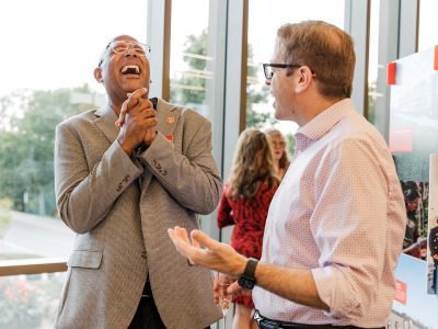 Two men stand in conversation, one throws his head back in laughter.