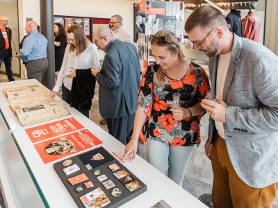 People holding glasses of wine and champagne look down at displays of printed materials and artifacts in a bright indoor space.