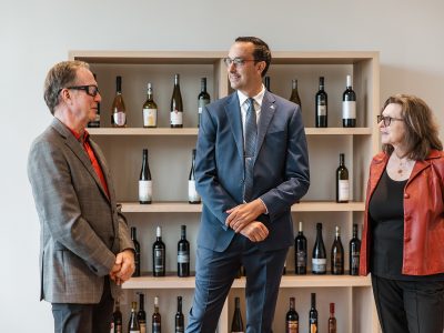 Three people stand in conversation in front of a bookcase displaying bottles of wine.