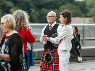 A man wearing a traditional kilt speaks with three women at an outdoor event.
