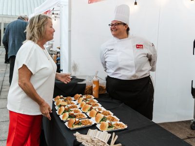 Two women, one wearing a chef's uniform, smile and chat over a table filled with plates of food.
