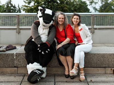 Two women pose for a photo sitting on a ledge outdoors with Brock University mascot Boomer the Badger.