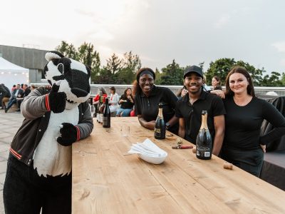 Three bartender's stand at a wooden counter with bottles of sparkling wine, posing for a photo with Brock University mascot Boomer the Badger.