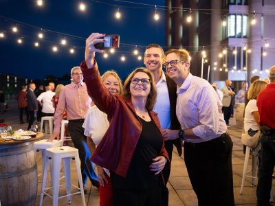 A woman wearing glasses holds up her phone to take a selfie with three other people at an evening rooftop party.