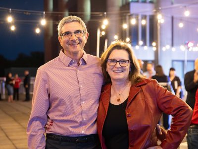 Two people with one arm around each other pose for a photo under strings of lights at an evening party outdoors.