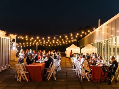 Guests dine at two long tables under twinkle lights at an outdoor evening party.