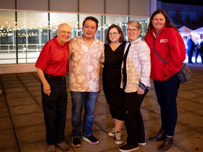 Five people pose for a group photo outdoors at a party during the evening in front of brightly lit windows.