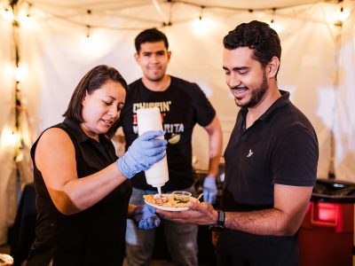 A man smiles holding a plate of food as a woman wearing rubber gloves squeezes a condiment bottle over the food.