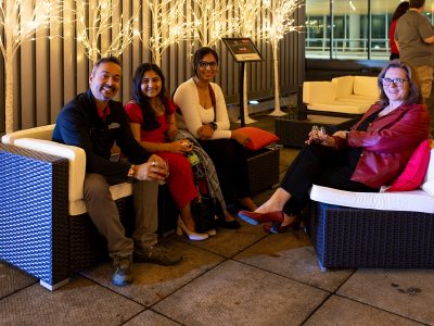 Four people sitting on patio furniture smile for a photo, a row of trees with twinkle lights behind them.