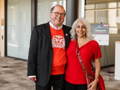 A man and a woman, each with an arm around the other, pose for a photo in a brightly lit room with windows in the background.