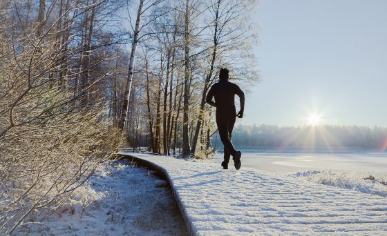 Young, adult alone man in tracksuit and hat running on forest trail in sunny winter morning after first snow.