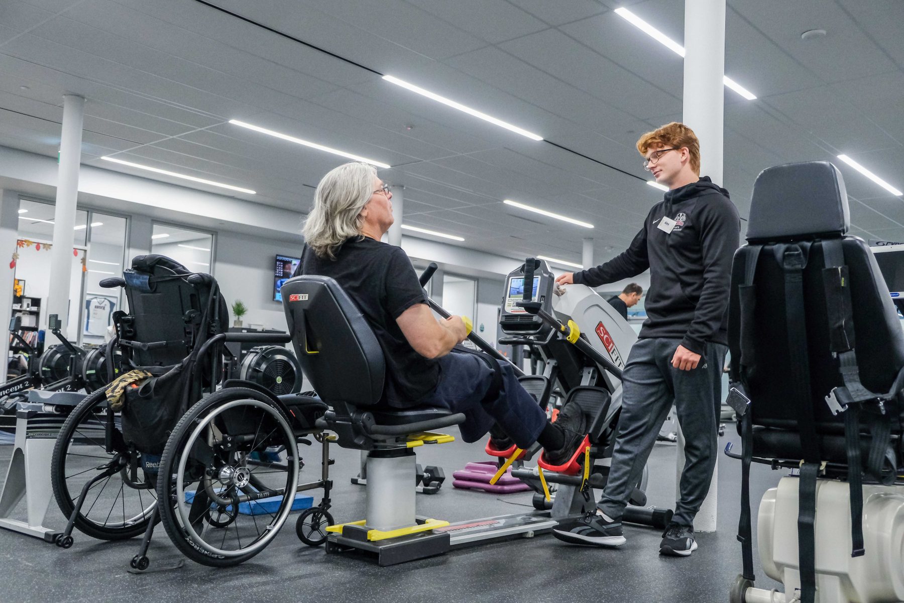 A university student speaks to a man using an exercise bike. His wheelchair is parked next to him.