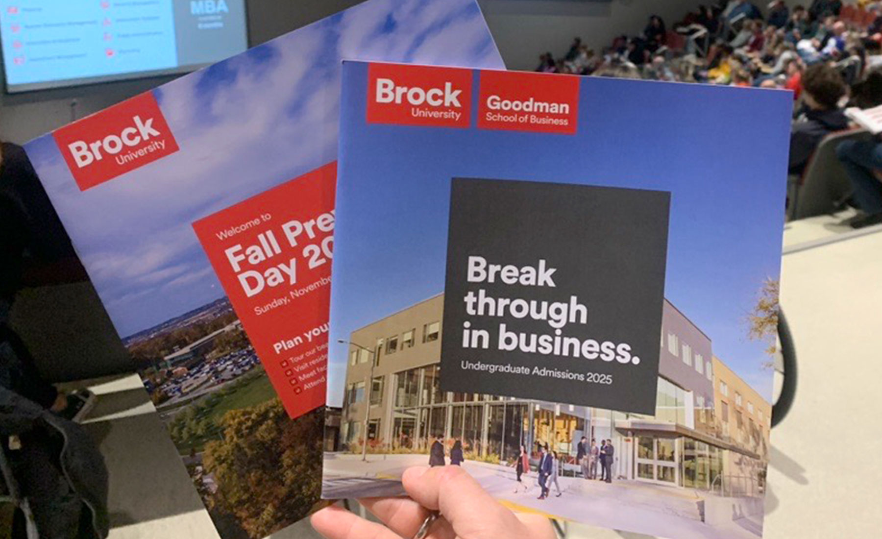 visual-elements2 A person holds two Brock University brochures in a lecture hall, with students seated and a presentation screen in the background.