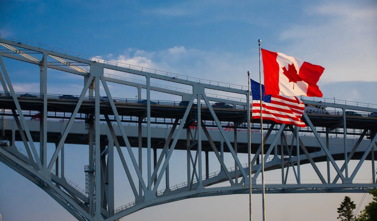 A metal bridge crossing with the U.S. and Canada flags flying from flagpoles in the foreground.