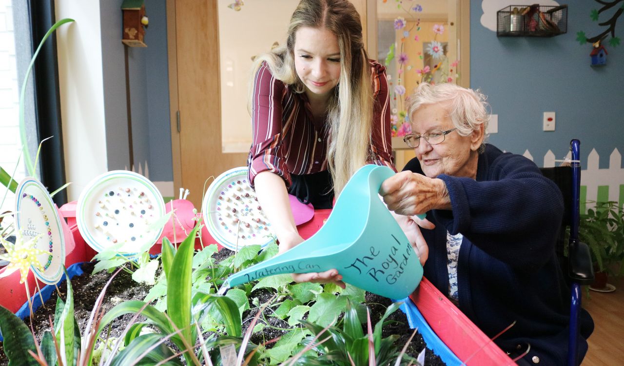 An elderly woman uses a watering can to water plans while a younger woman assists.