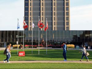 Three people walk on a sidewalk in front of a series of flags and a large building with a tall concrete tower in the background.