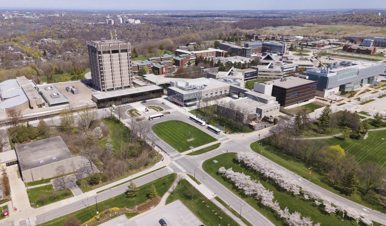An aerial photograph of Brock University’s St. Catharines campus, which is made up of several buildings of various sizes, asphalt parking lots and roadways, and plenty of greenspace with trees and grass.