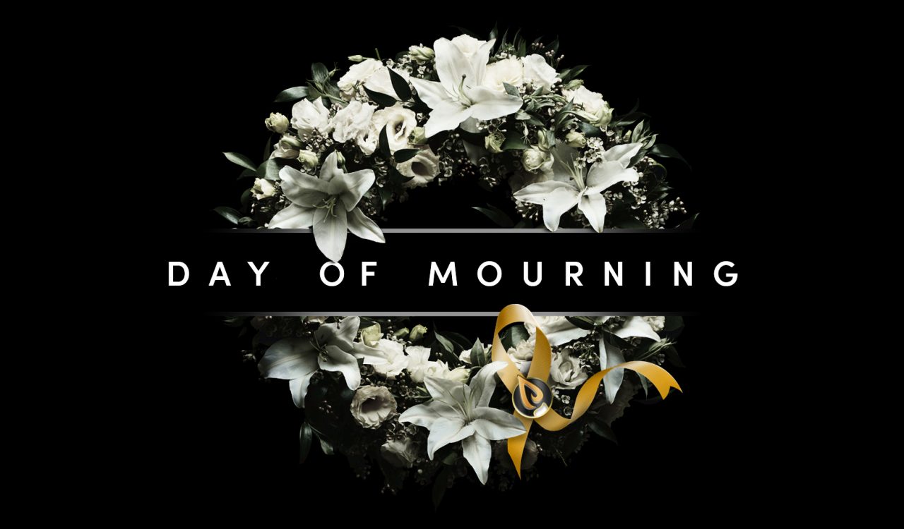 A wreath with white flowers and a yellow ribbon has the words “Day of Mourning” written across it.