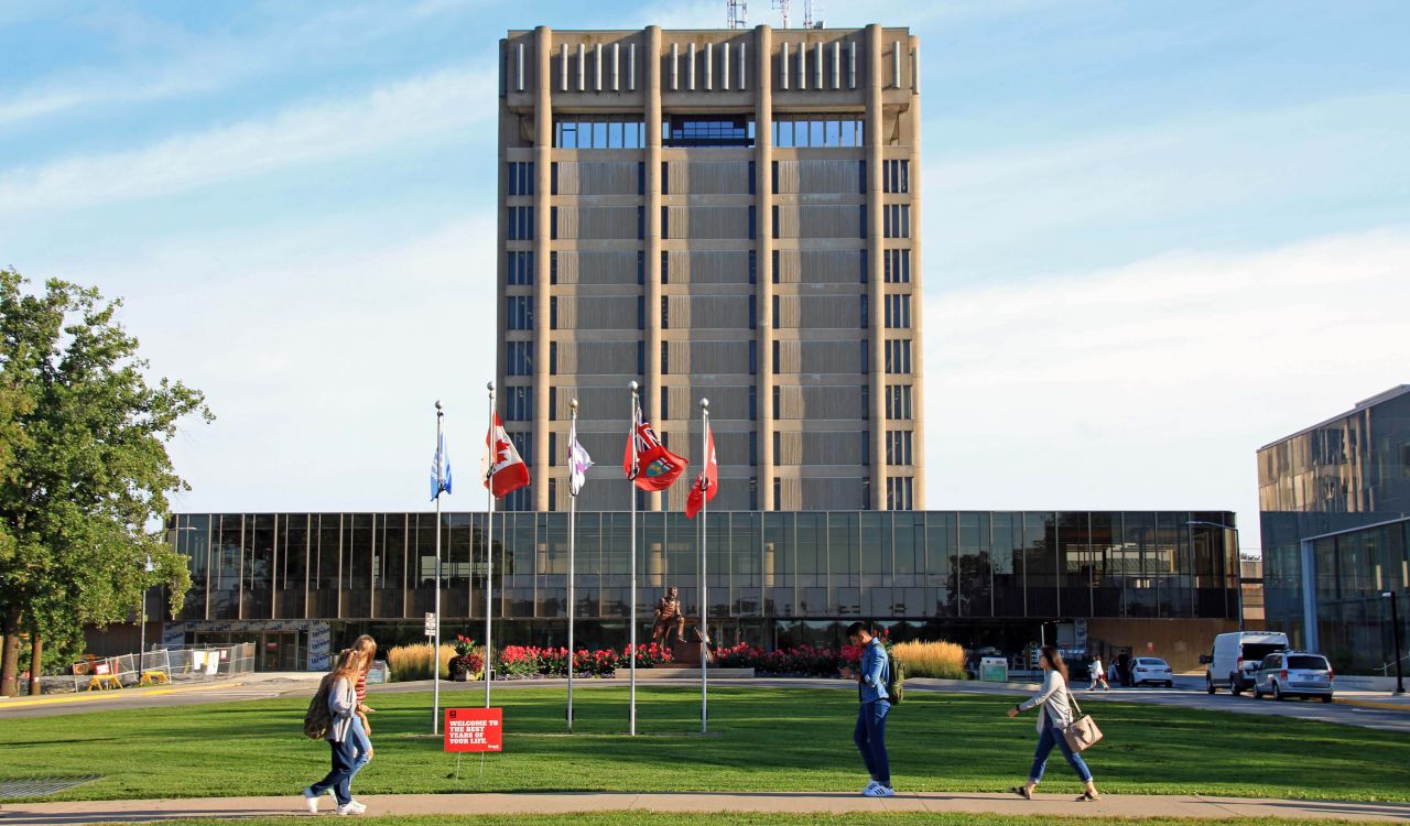 A 13-storey brown building against a blue sky in the background, with students walking on a sidewalk surrounded by green space in the foreground.