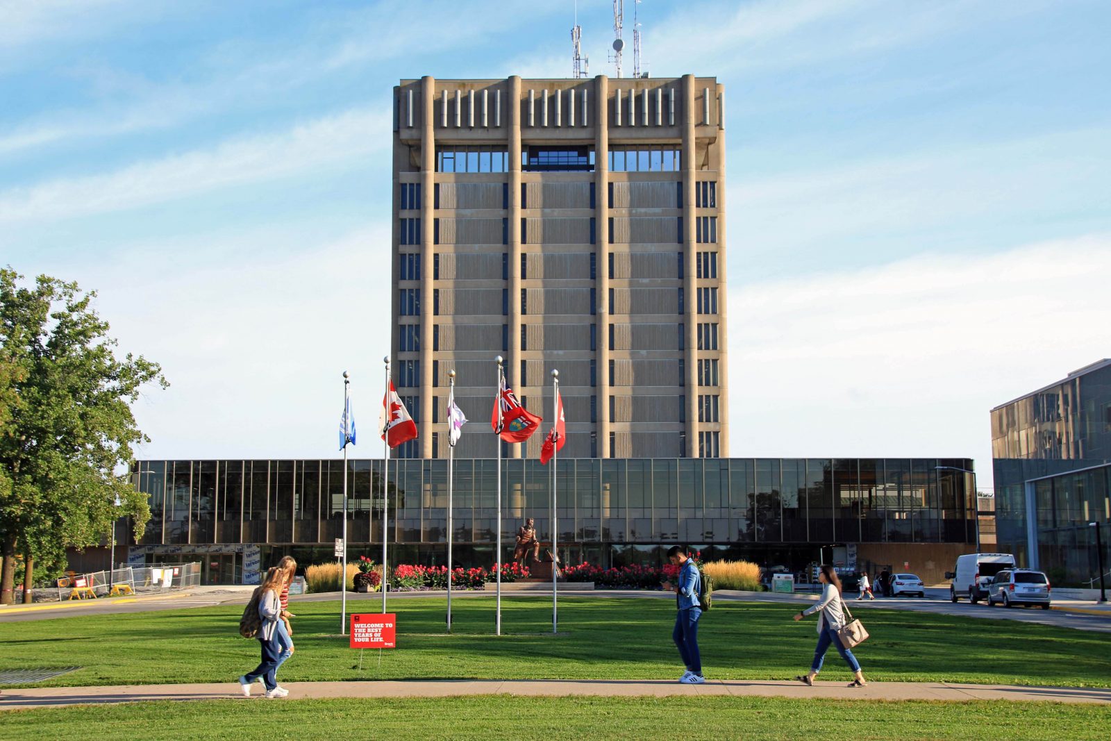 A 13-storey brown building against a blue sky in the background, with students walking on a sidewalk surrounded by green space in the foreground.