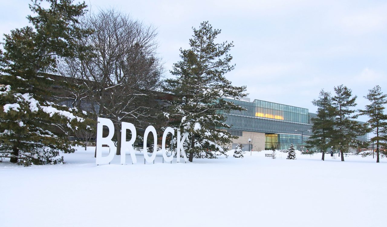 Giant letters spelling out "Brock" stand in front of trees in a snowy field with a glass building in the background.