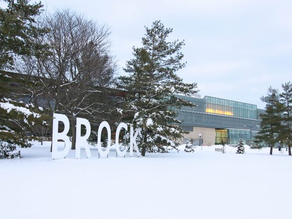 Giant letters spelling out "Brock" stand in front of trees in a snowy field with a glass building in the background.