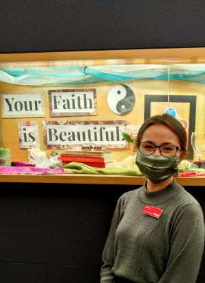 A young masked woman stands in front of a display that says "Your Faith is Beautiful."