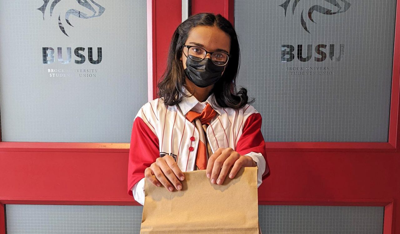 A young masked woman holds a brown paper bag in front of the doors to the Brock University Students' Union offices