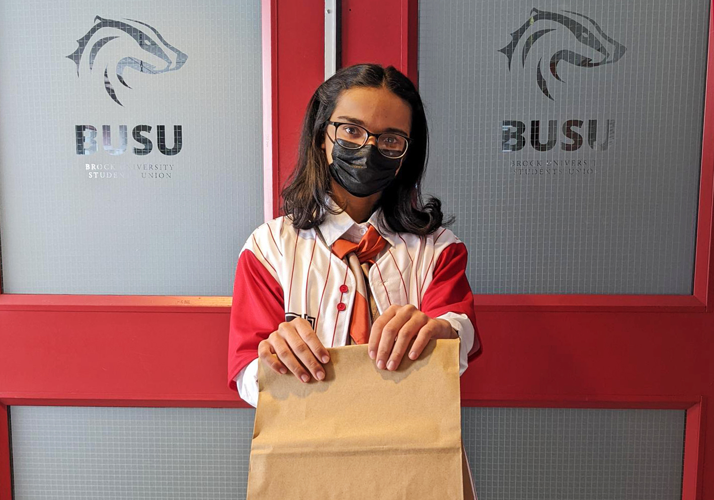 A young masked woman holds a brown paper bag in front of the doors to the Brock University Students' Union offices