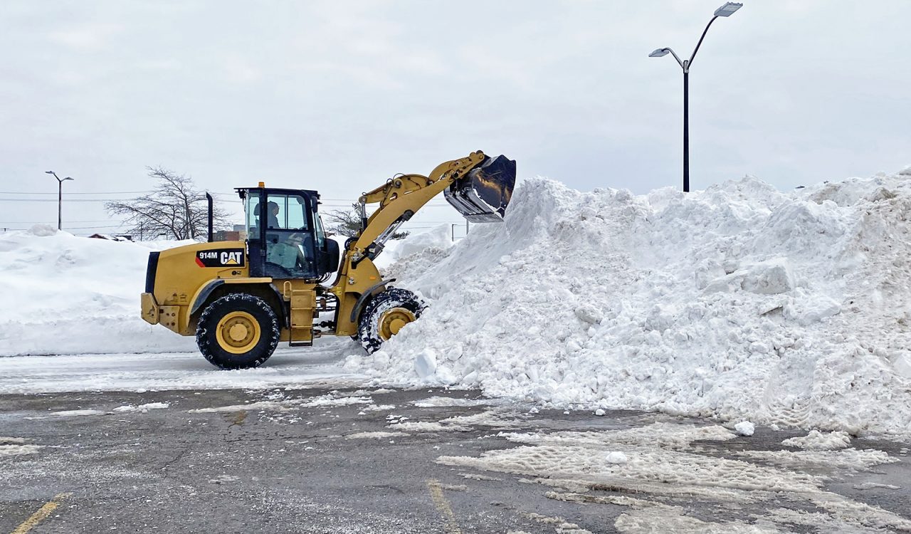 A horticultural technician uses a yellow wheel loader to scoop snow onto a large snow pile in a parking lot at Brock University.