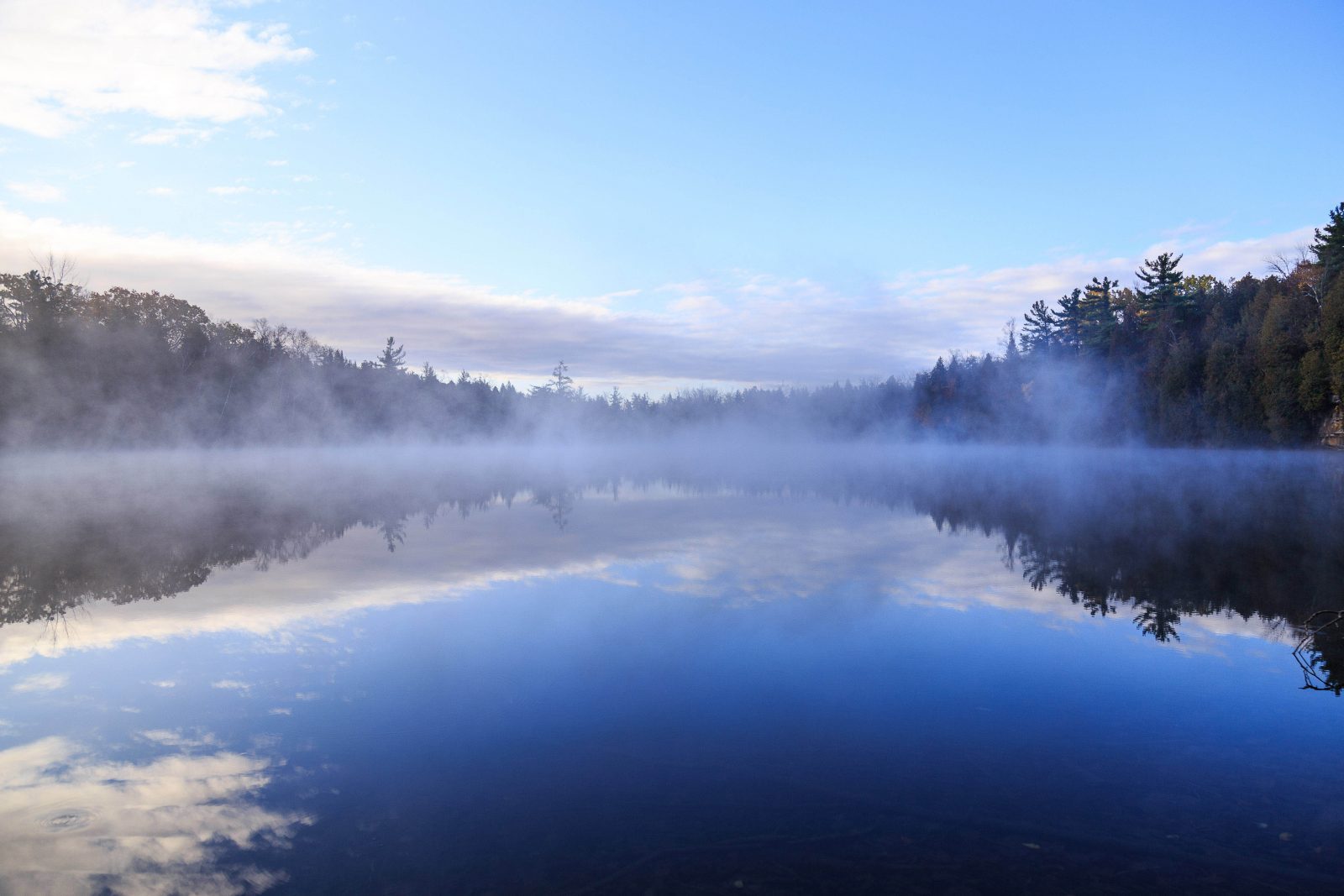 Fog rising over a lake with a treeline and blue sky in the background