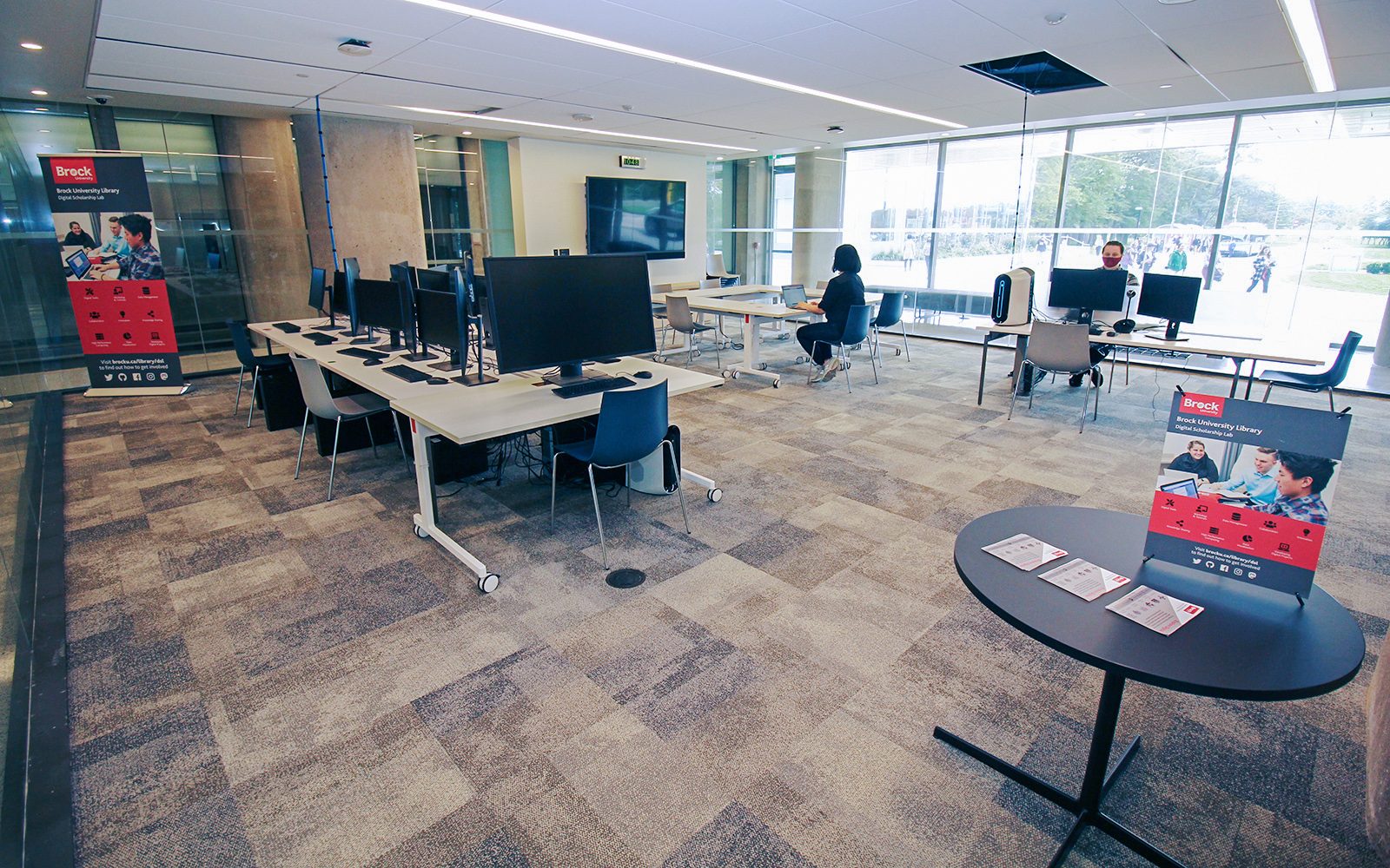 A large glass enclosed room contains several computers on white tables and a large digital screen on one wall. Two people sit at tables: one uses a laptop, the other uses one of the desktop computers.