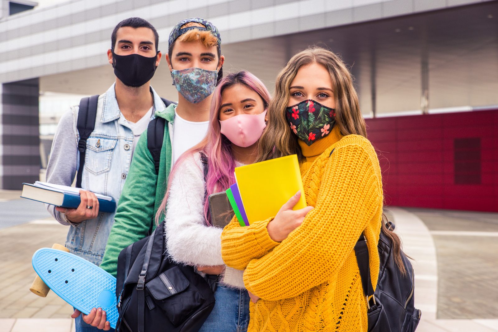 Four masked teenagers carrying books and a skateboard stand together in front of a building