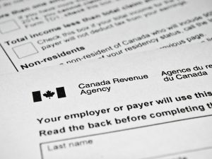 A closeup photo of two tax forms from Canada Revenue agency. A black and white Canada flag is in the top left corner of the top form. Below the flag are blocks of texts and open boxes to fill in information.