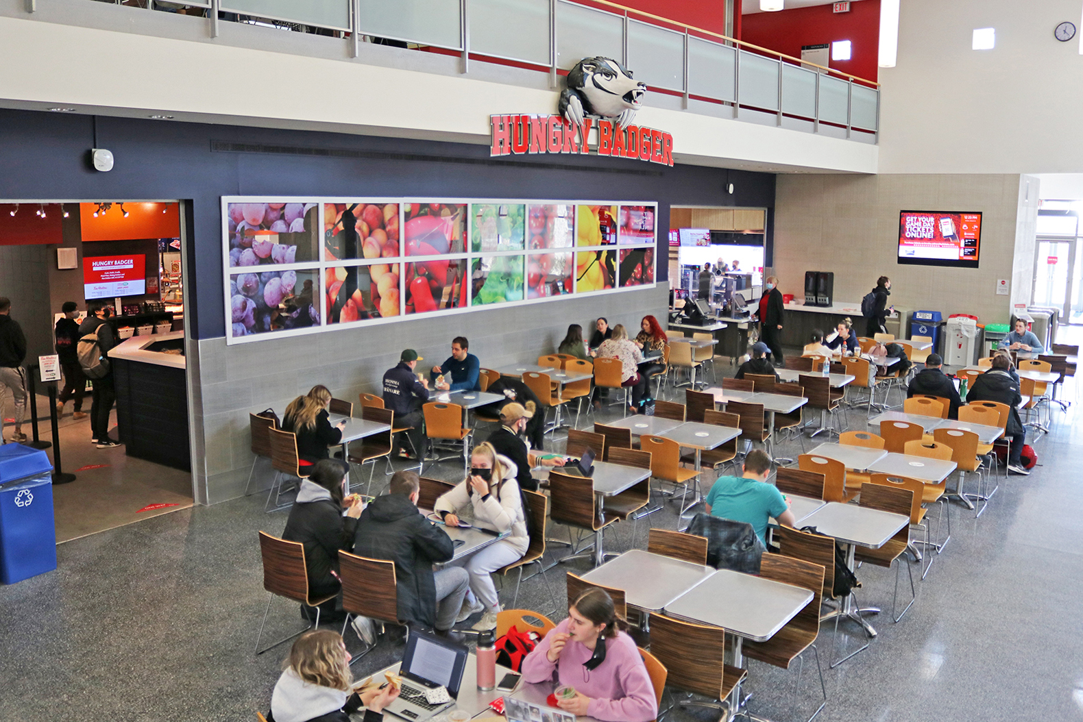 Several people sit at tables and chairs while eating lunch and working on laptops. A wall behind them features several large photos of colourful fruits and vegetables. Above the photos is a sign with the words “Hungry Badger” in capital letters and a cartoon 3D image of a badger.