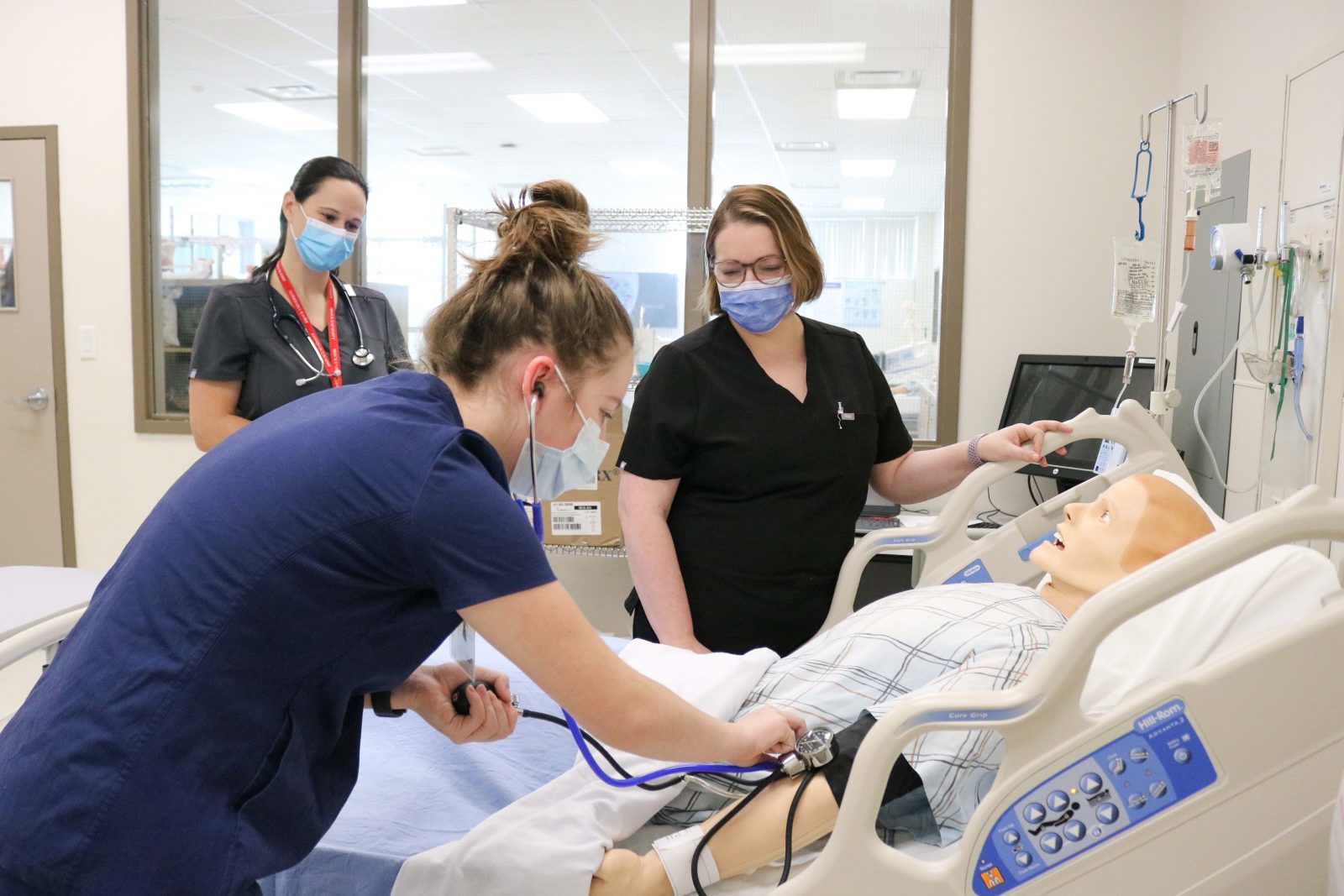 A female Nursing student checks the blood pressure of a simulator that looks like a male patient as two supervisors look on