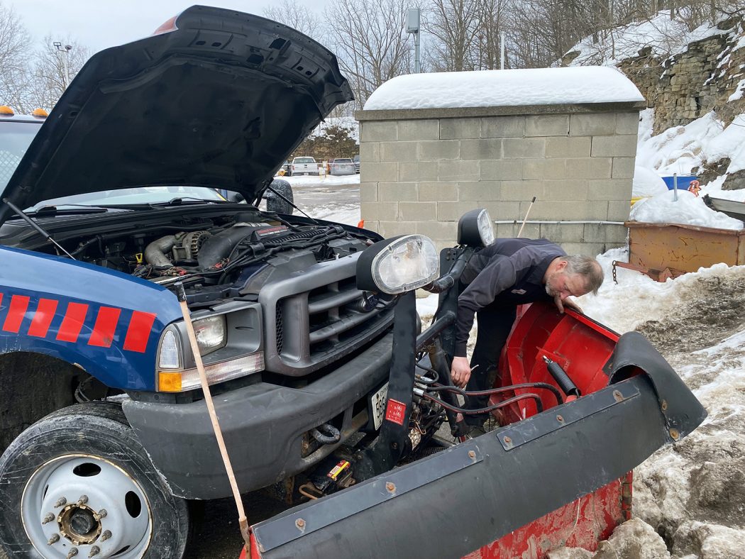 A mechanic bends over to work on the electrical components of a snowplow that is attached to the front of a pickup truck.