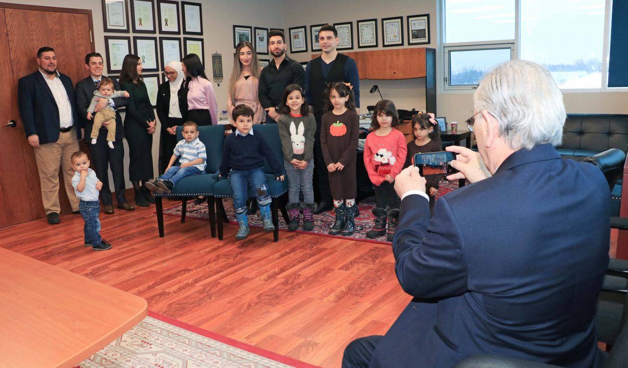A man holds up his phone to take a photo of eights adults and eight children in the corner of an office.
