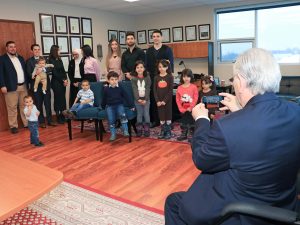 A man holds up his phone to take a photo of eights adults and eight children in the corner of an office.