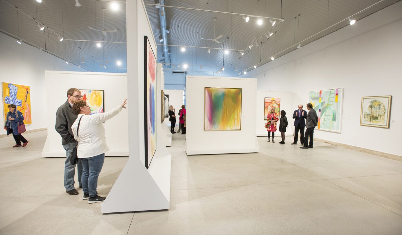 Small groups of people stand in a bright, white room looking at colourful paintings on the wall. The square and rectangular paintings are abstract images using bright yellow, green and pink colours. Track lighting hangs from the ceiling.