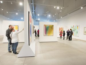 Small groups of people stand in a bright, white room looking at colourful paintings on the wall. The square and rectangular paintings are abstract images using bright yellow, green and pink colours. Track lighting hangs from the ceiling.