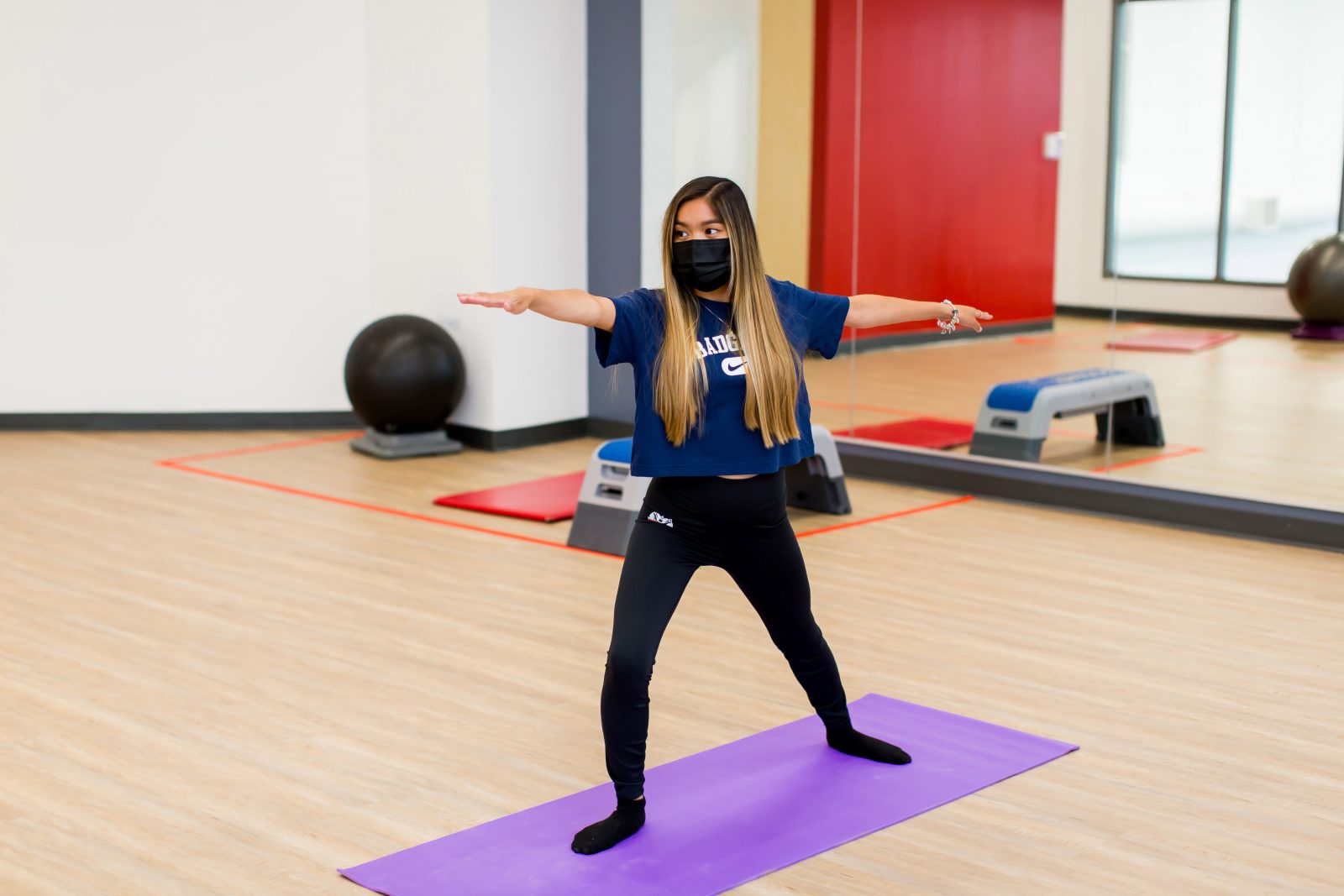 A masked woman stands on a purple yoga mat in a gym with her arms outstretched. She has waist-length hair and is wearing a black face mask, dark blue Nike T-shirt with ‘Badgers’ written on it, black yoga tights and black socks.