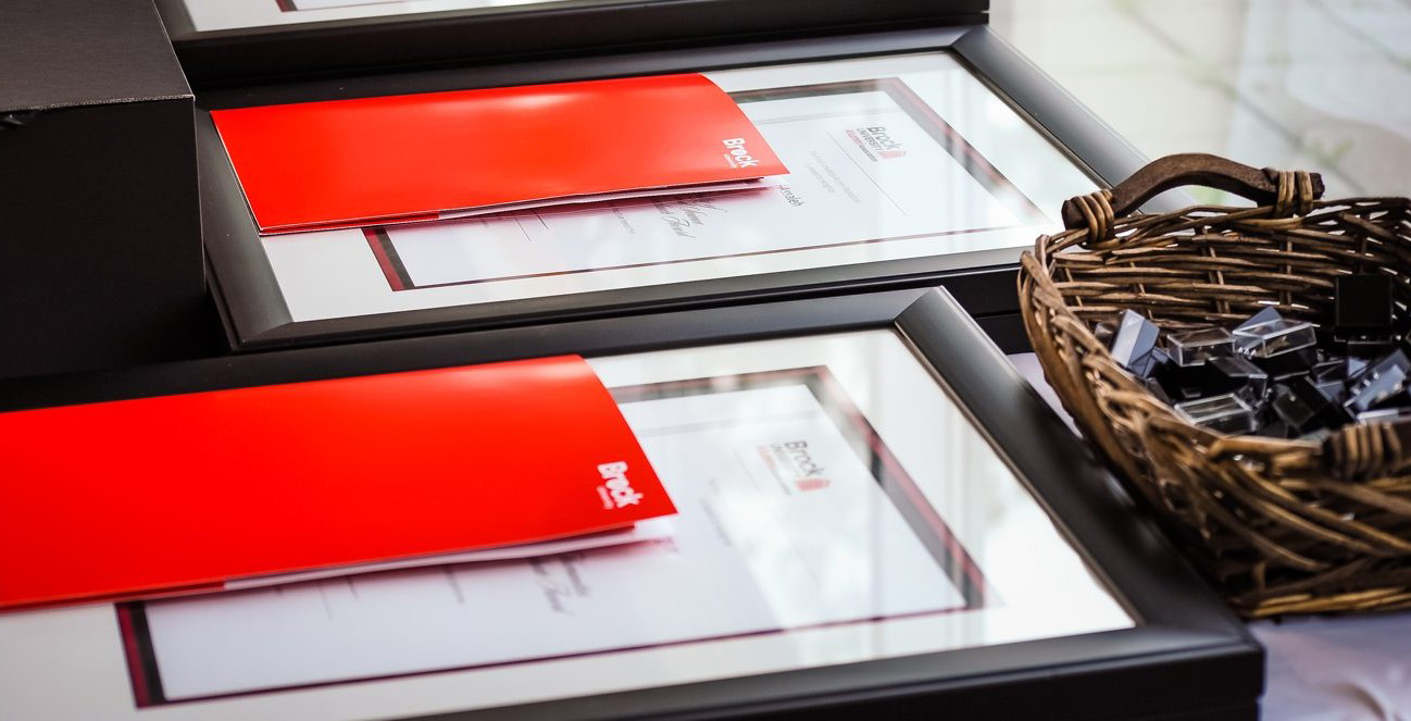 Two framed award certificates and red file folders rest on top of a table beside a basket of lapel pins.