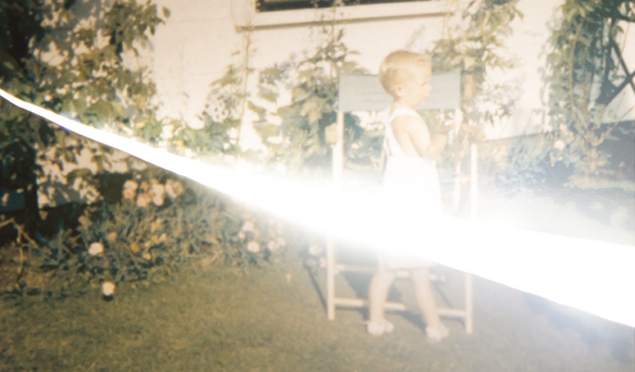 A slightly blurred photograph of a small child dressed in white shorts standing in front of a chair, outside a white house with a small black window in the background. The child is standing on green grass with green plants lining the house, and a streak of white resembling light dramatically strikes through the centre of the photo.