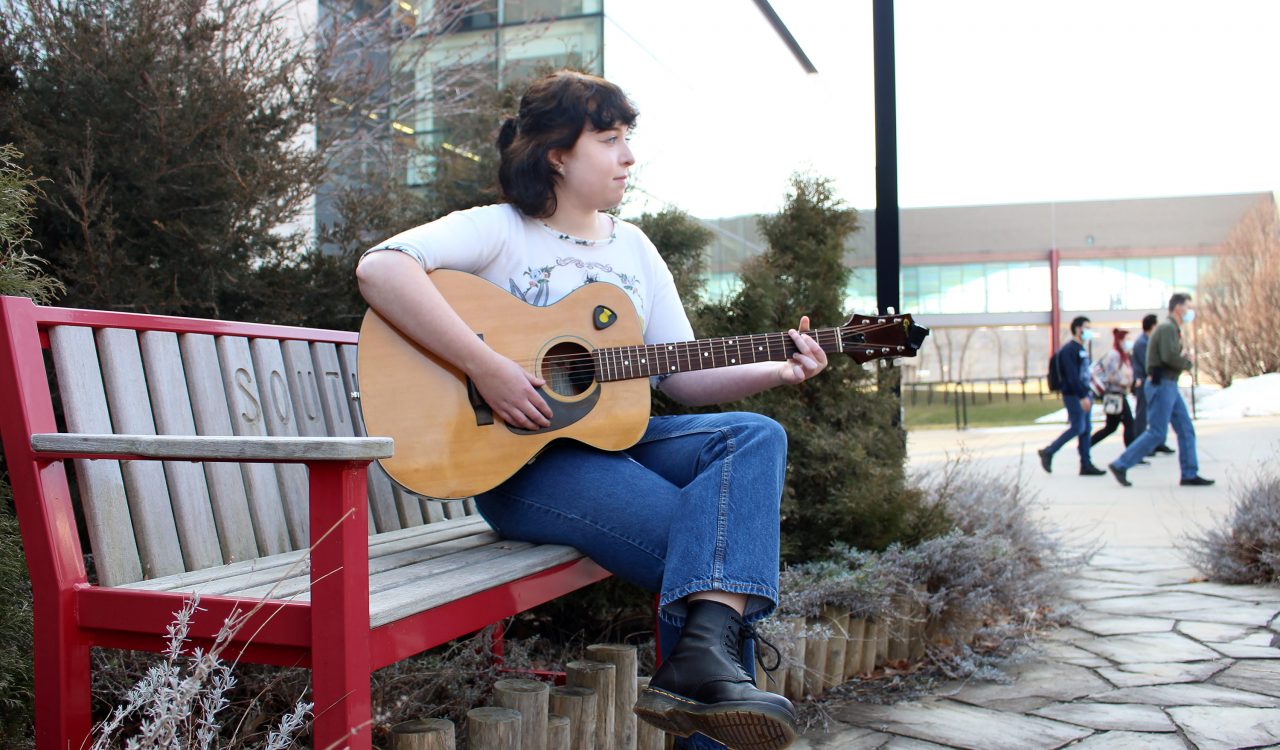 A woman is pictured sitting on a bench strumming a guitar surrounded by plants in front of a glass building on Brock’s main campus. A group of students walk past in the background.