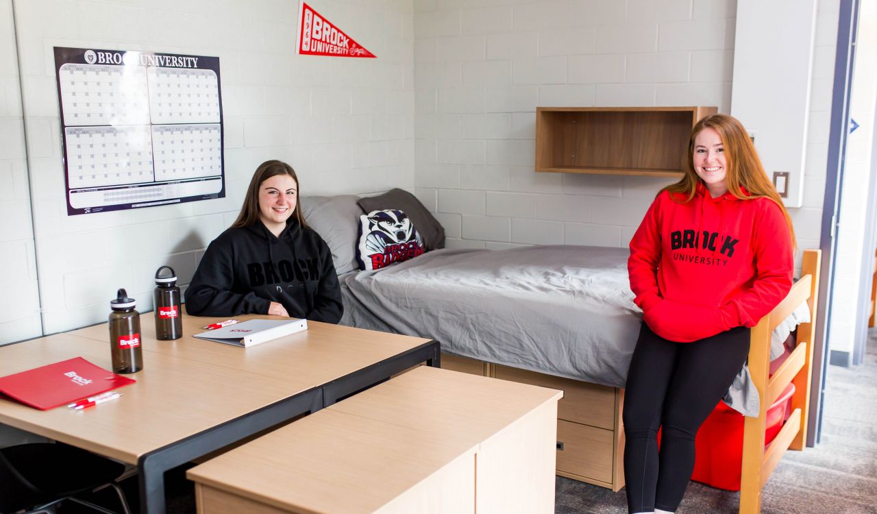 Two female roommates in a university dorm room. One is seated behind a desk and the other is leaning against the bed.