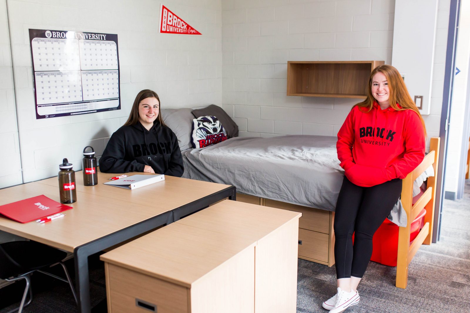 Two female roommates in a university dorm room. One is seated behind a desk and the other is leaning against the bed.
