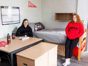 Two female roommates in a university dorm room. One is seated behind a desk and the other is leaning against the bed.